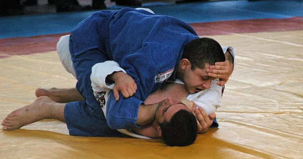 BJJ competitor in closed guard preparing a triangle choke while a judo player in gi attempts to posture up — illustrating the fundamental split between the two arts: judo prioritizes standing throws, BJJ prioritizes ground submissions.