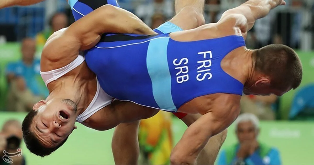 Dois lutadores competindo em um tatame olímpico — um com collant azul, outro com vermelho, ilustrando o combate em clinch focado na parte superior do corpo que define o estilo greco-romano. Na luta livre, as pernas são um alvo legal; na greco-romana, estão completamente proibidas.