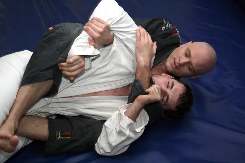 Back control with hooks in during BJJ training — the attacker has the seatbelt grip and is setting up the rear naked choke. Public domain via Wikimedia Commons